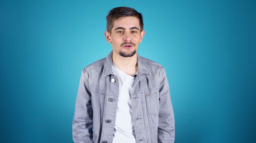 A young white male student, wearing a white t-shirt and denim jacket, standing in front of a blue background