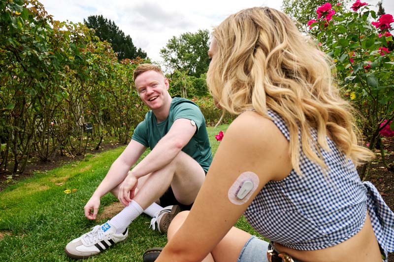 Two young people sat in a park, with type 1 diabetes continuous glucose monitor visible on their arms