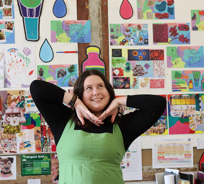 Megan Neville standing in her bright and colourful university workspace. The wall behind her is covered in bright and colourful art work. Megan poses playfully smiles and looks to the side with her hands under her face.