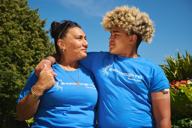 A woman and a young man looking at each other while standing outside in the sun. They are wearing T-shirts to represent Breakthrough T1D, a funder of type 1 diabetes research.