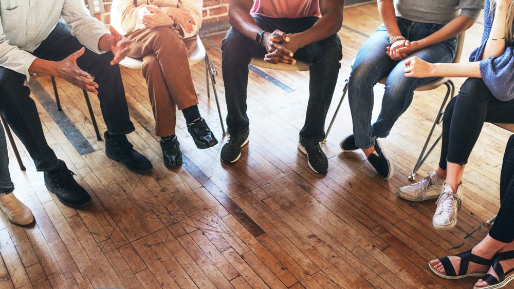 Anonymous circle of people sat in chairs, shown from waist down, in a communal space.