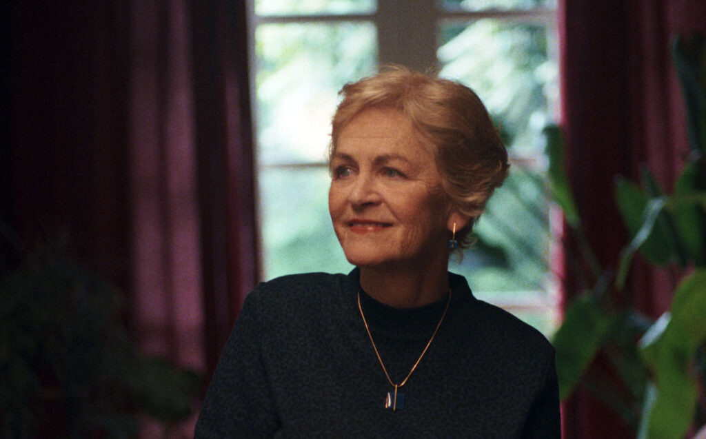 Liz sat at dining table smiling, from the film What a Cure Feels Like
