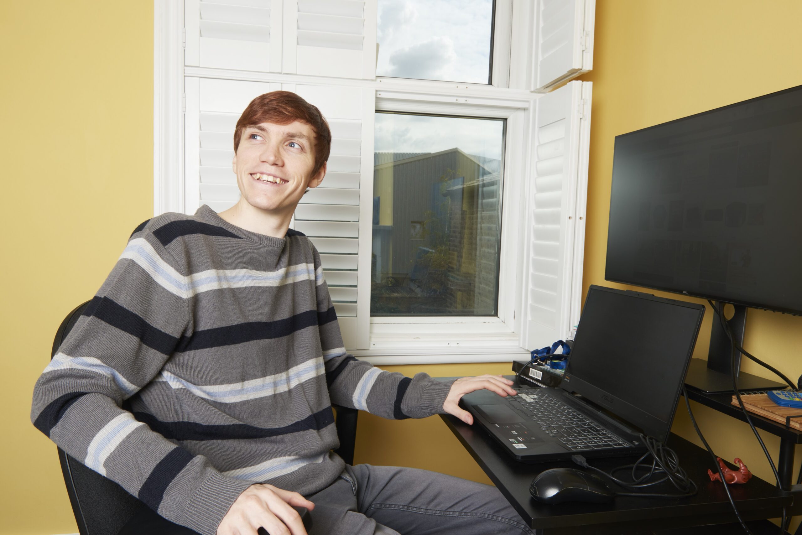 A young man is sitting at a desk with a computer, looking over his shoulder and smiling.