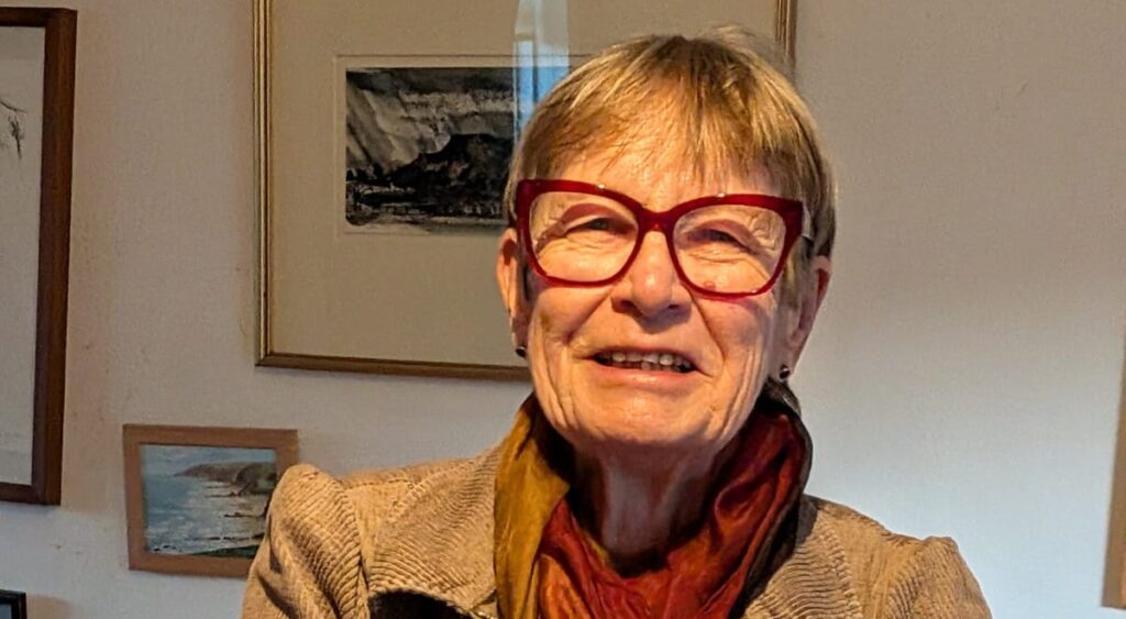 A head and shoulders photograph of an older woman wearing red glasses and smiling at the camera