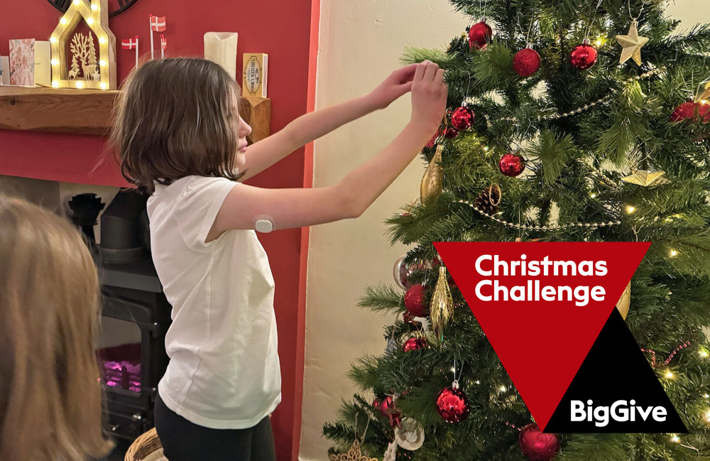 Girl with CGM on arm decorating a Christmas Tree, with Big Give Christmas Challenge logo