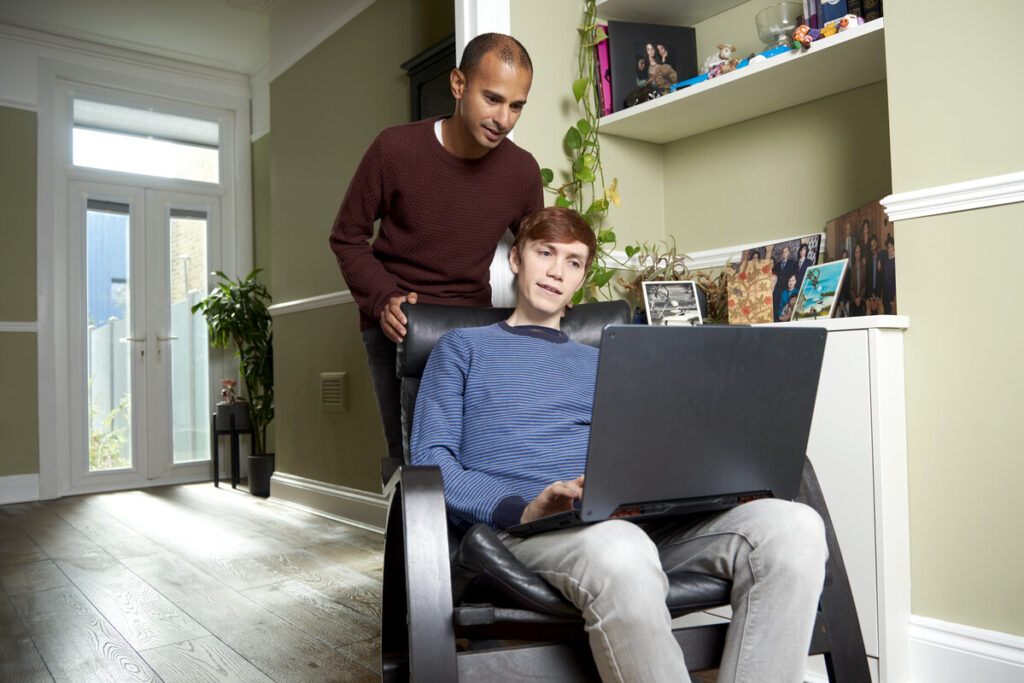 Man in blue jumper sat on chair looking at computer. Man in purple jumper looking over his shoulder