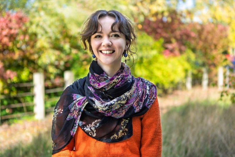 A woman standing outside with greenery behind her, smiling and staring into the camera