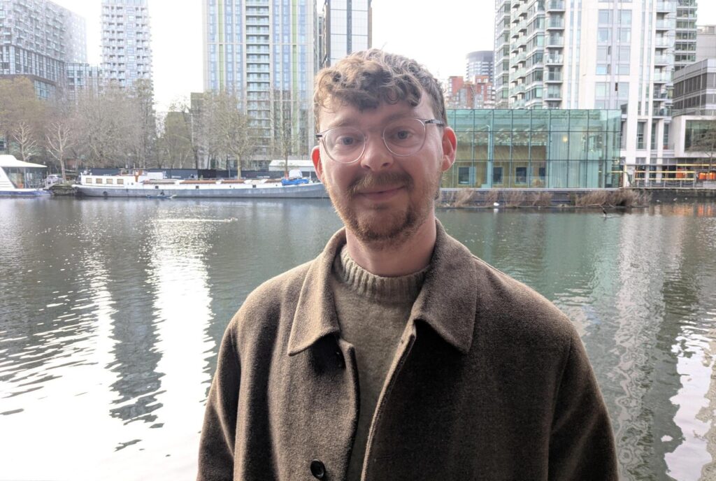A man in a brown jumper and brown coat staring into the camera