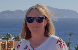 A woman wearing sunglasses looking towards the camera with the sea and mountains behind her.