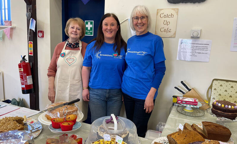 Bake sale stall with various cakes and people in Breakthrough T1D shirts with "Catch Up Cafe" sign behind them