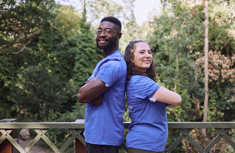 Two smiling adults stood back to back in blue Breakthrough T-shirts in the park, CGMs visible on folded arms.