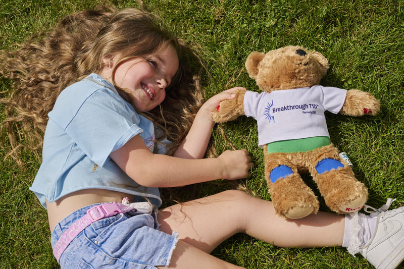 Smiling girl lying next to Breakthrough T1D Rufus bear in park