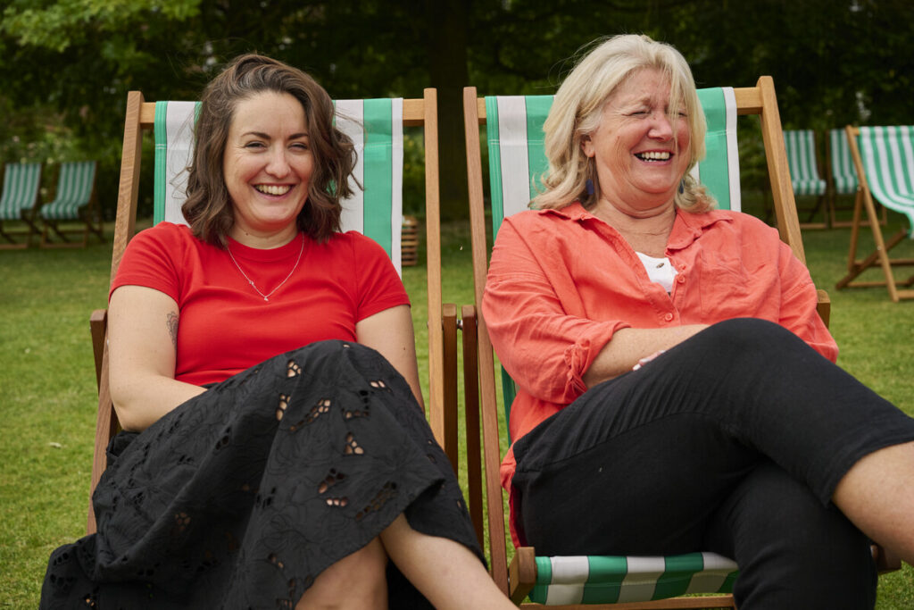 Two women wearing red tops, sat in deckchairs in the park, laughing.